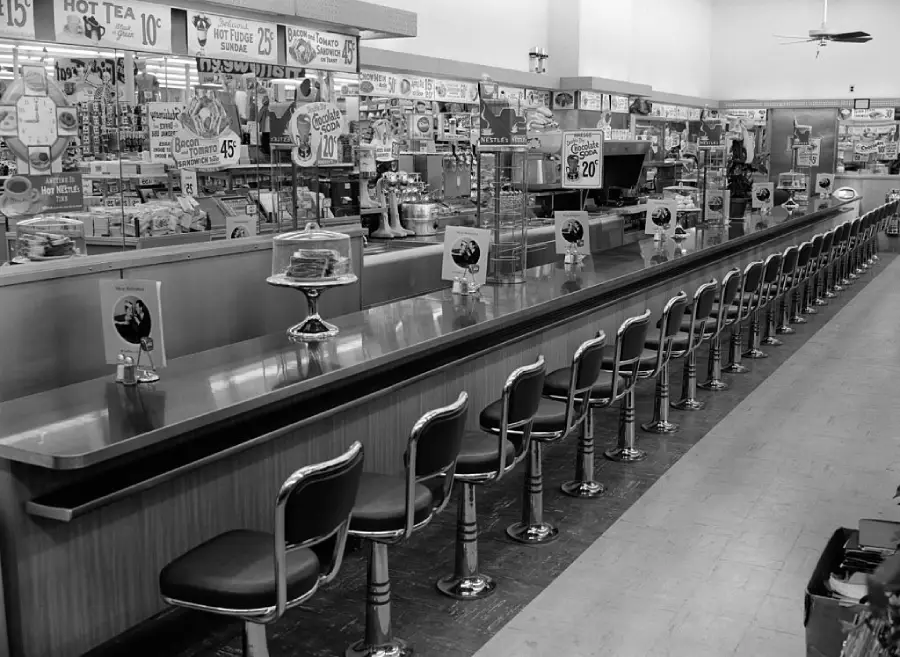 A long counter with stools in a retro diner or lunch counter, with menus and cake displays. Shelves behind the counter hold various products and signs advertise drinks and food specials.