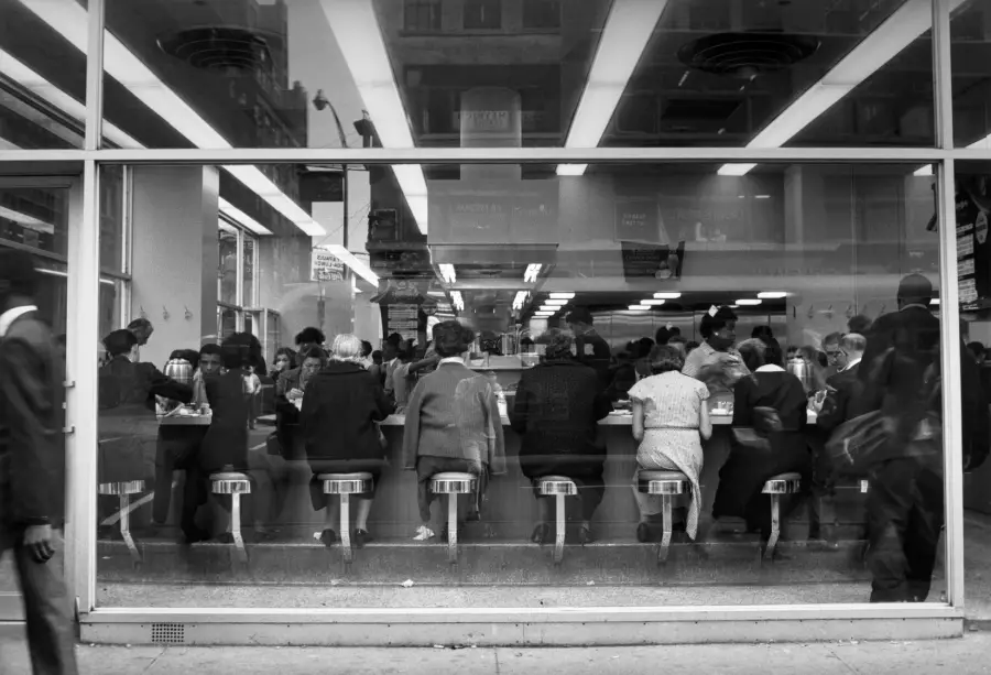 Black and white photo of people sitting at a counter inside a diner, viewed from the street through large glass windows. Some are eating or talking; stools line the counter. The scene looks busy and lively.