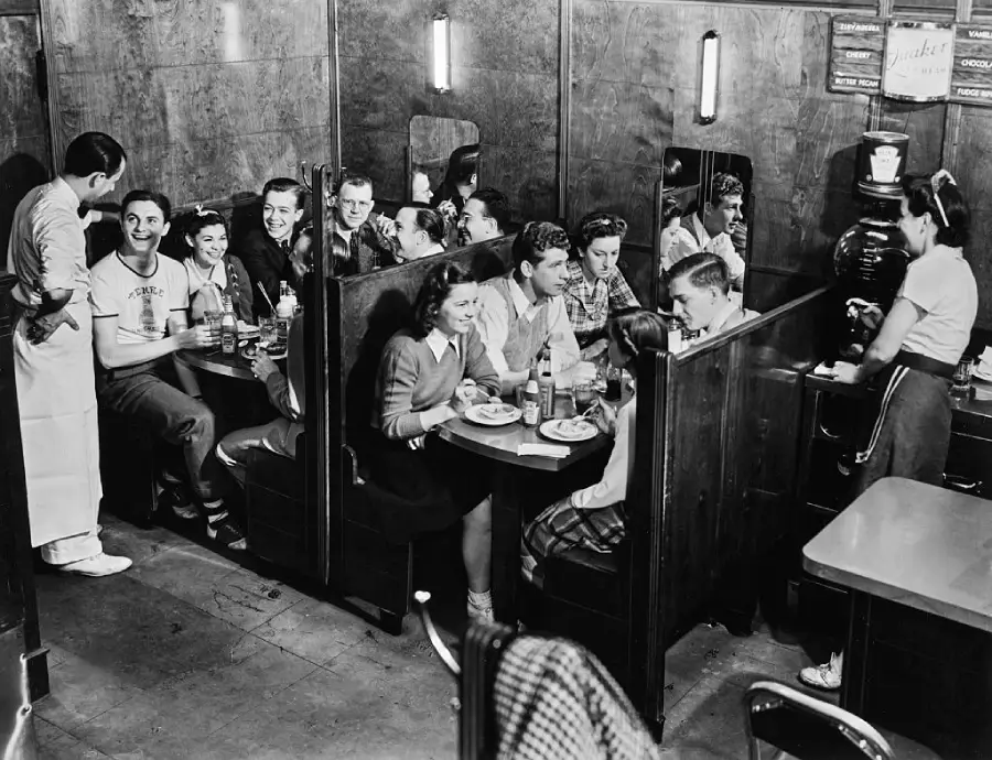 Black and white photo of teens sitting in diner booths, eating and drinking, while a waitress serves coffee and a cook stands nearby, smiling and talking to them. The scene feels lively and social.