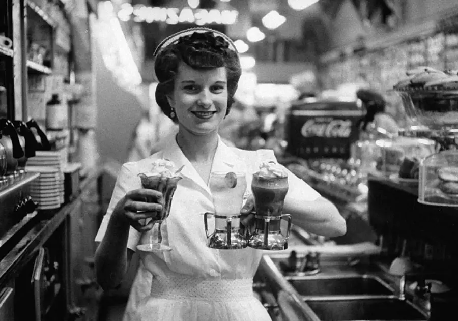 A smiling waitress in a 1950s diner holds two ice cream sundaes. She wears a white uniform and stands behind the counter, with various diner items and a Coca-Cola fountain machine in the background.