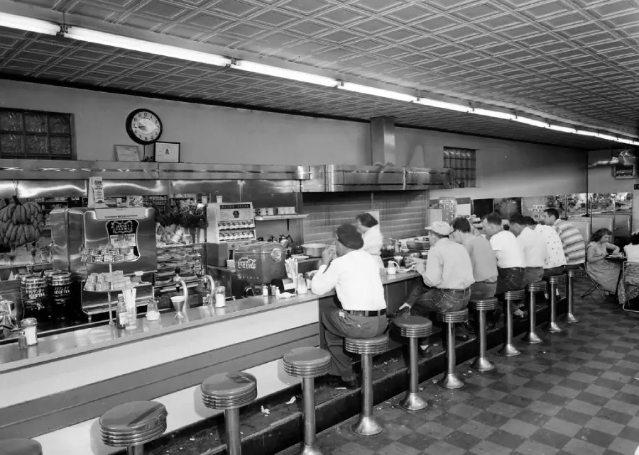 Black-and-white photo of a vintage diner counter with people sitting on stools, eating and drinking. Soda machines, cups, and menus are visible behind the counter. The scene has a classic 1950s atmosphere.