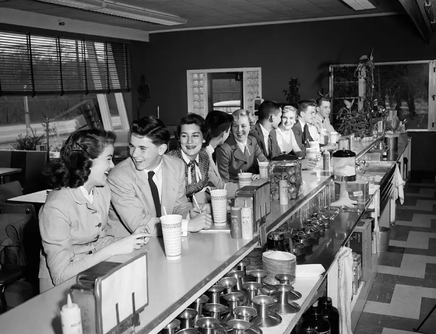 A group of young people in 1950s attire sit at a long diner counter, smiling and chatting while holding drinks. The diner has checkered floors and various containers and appliances on the counter.