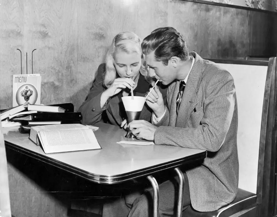 A man and woman sit together in a booth, both sipping from the same milkshake with straws. An open book and a menu are on the table, and they appear to be enjoying each other’s company.