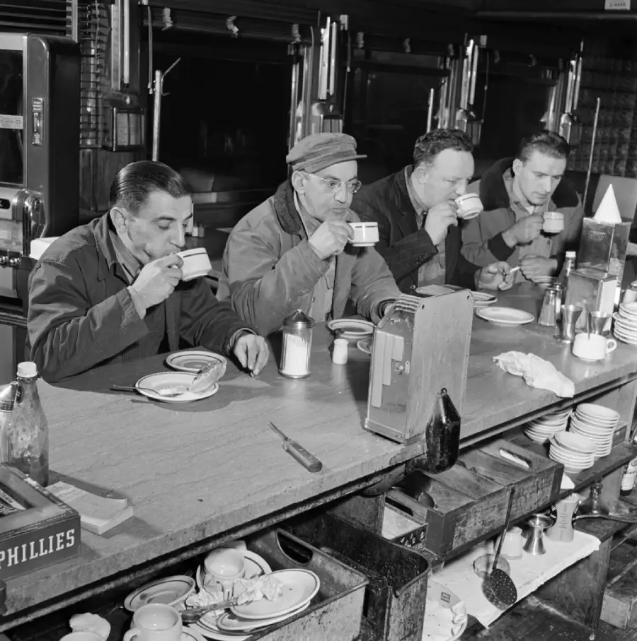 Four men sit side by side at a diner counter, drinking from cups. Plates, utensils, a napkin holder, and condiments are on the counter. The scene appears to be from the mid-20th century.
