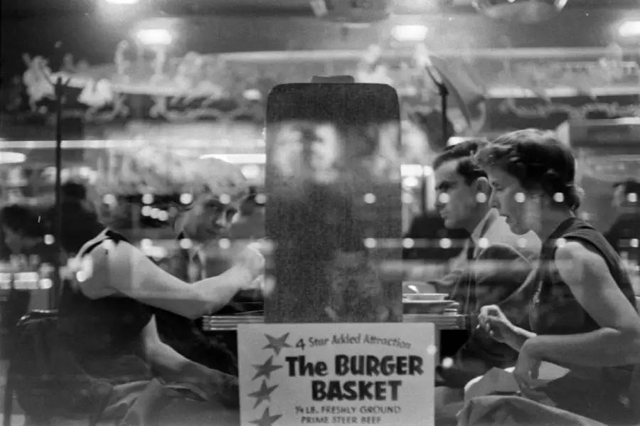Three people sit at a diner booth, seen through a window with reflections. A sign on the window advertises “The Burger Basket.” The scene appears to be from a past era, possibly the 1950s or 1960s.