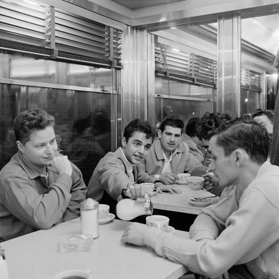 Four young men sit at a diner booth, drinking coffee. One pours cream into another’s cup. They appear relaxed and engaged in conversation. The diner has metallic walls and large windows reflecting the interior lights.