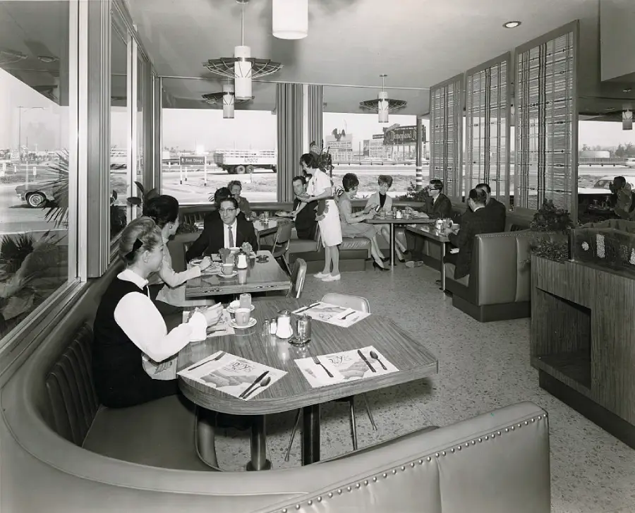 Black-and-white photo of a busy diner with people sitting in booths, eating and conversing. Waitresses serve food. Large windows show parked cars outside. Retro 1960s decor and furnishings are visible.