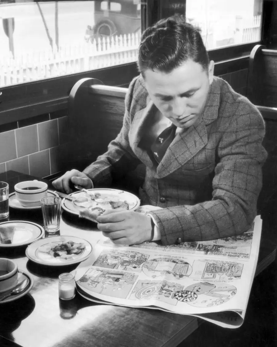 A man in a suit sits at a diner booth, eating breakfast while reading a large comic section of a newspaper. There are plates, coffee, and a glass of water on the table.