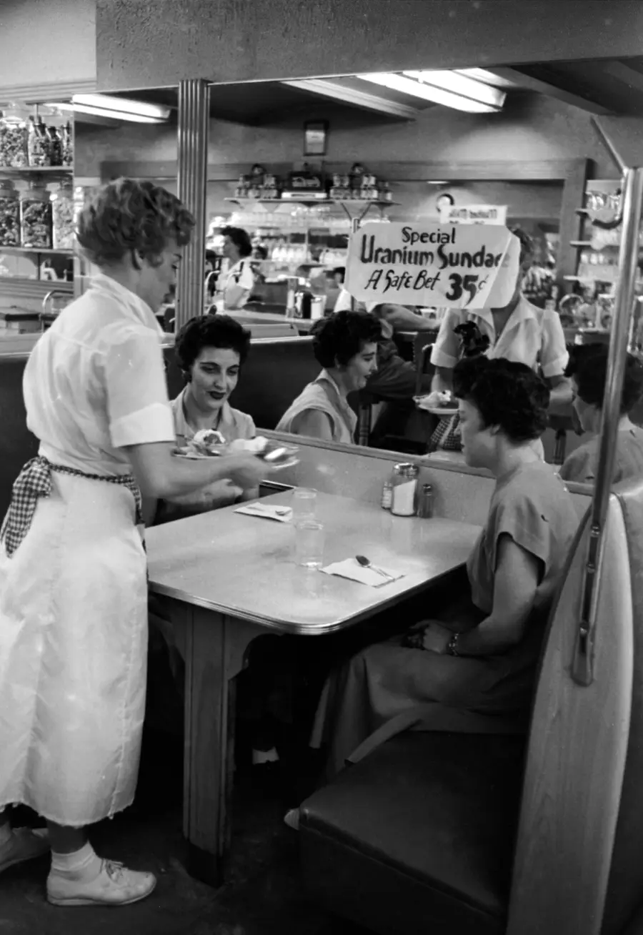A waitress serves ice cream to women at a diner booth. A sign in the background reads, “Special Uranium Sundae, A Split-Det 35¢.” Shelves of products are visible behind the counter.