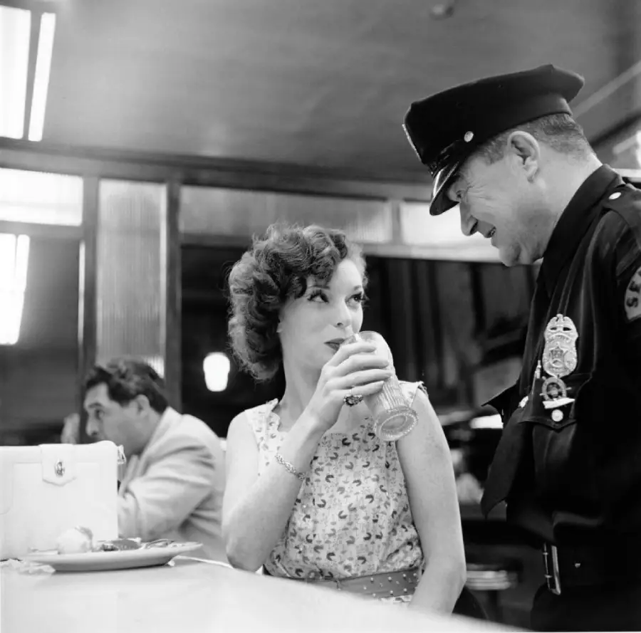 A woman in a sleeveless dress drinks from a glass at a diner counter while smiling up at a uniformed police officer; another man sits in the background.