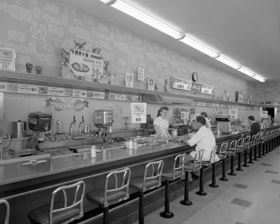 Black and white photo of a vintage diner counter with stools. Three workers in uniforms serve food, while two customers sit. Menu boards and food machines line the wall behind the counter.