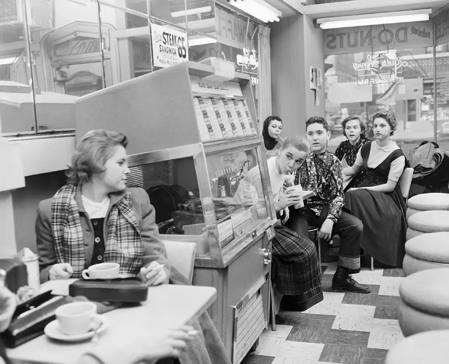 Black and white photo of young people in a diner, some sitting at a booth, others near a jukebox. Most are looking toward the camera. Coffee cups and trays are on the table. The setting appears to be from the 1950s.