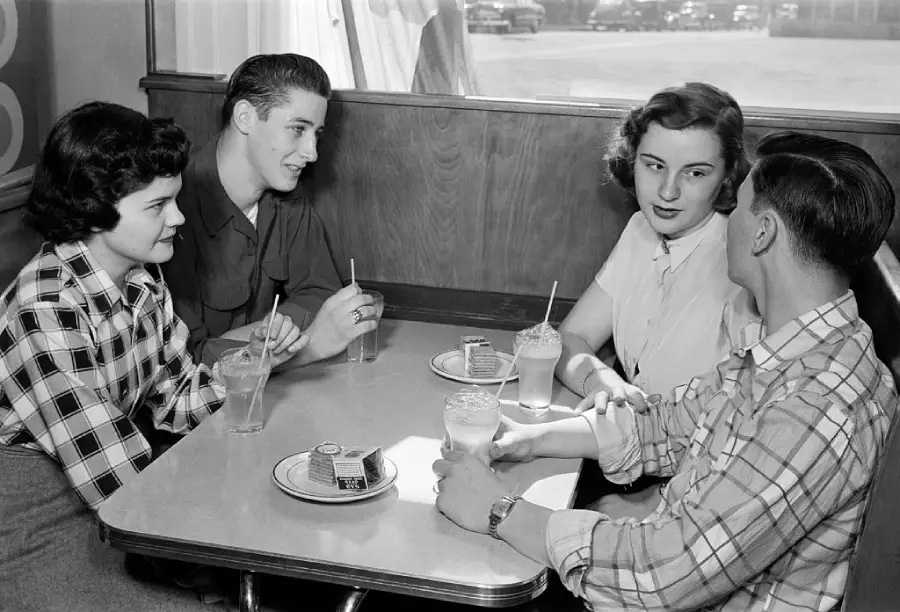 Four young adults sit at a diner table, drinking milkshakes and chatting. Two women and two men are seated across from each other, with plates of cake in front of them. The setting appears to be from the 1950s.