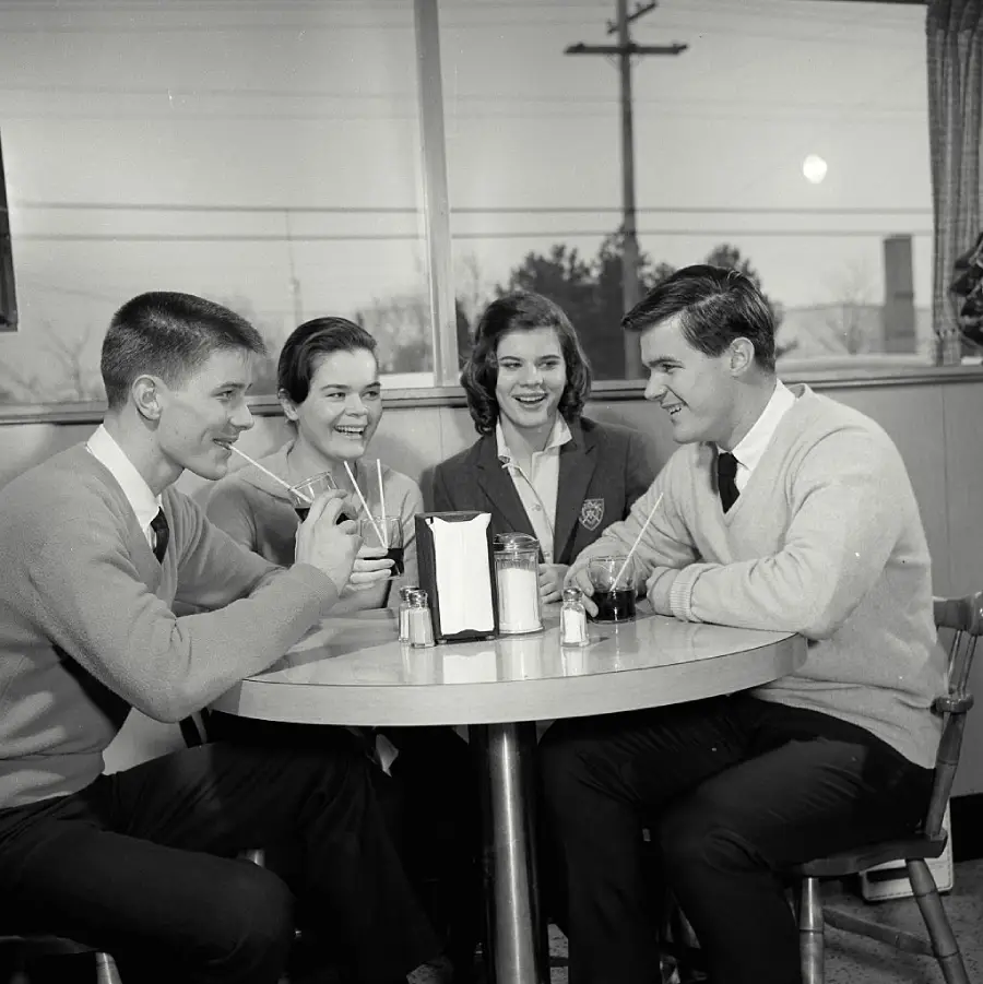 Four young adults in sweaters sit around a diner table, smiling and sharing drinks with straws. They appear to be enjoying a friendly conversation in a bright, casual setting.