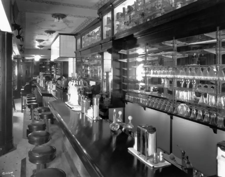 A vintage black and white photo of a long soda fountain bar with stools, glassware, and dispensers, reflecting an early 20th-century diner or pharmacy interior.