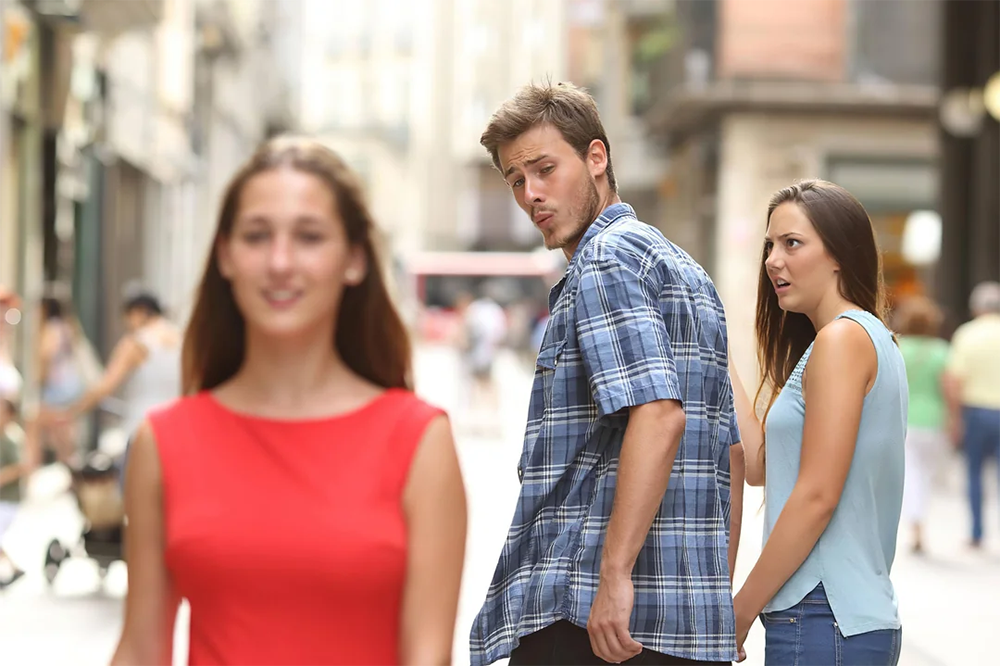 A man walking with a woman looks back at another woman in red passing by, while his companion looks at him with surprise and disbelief on a city street.