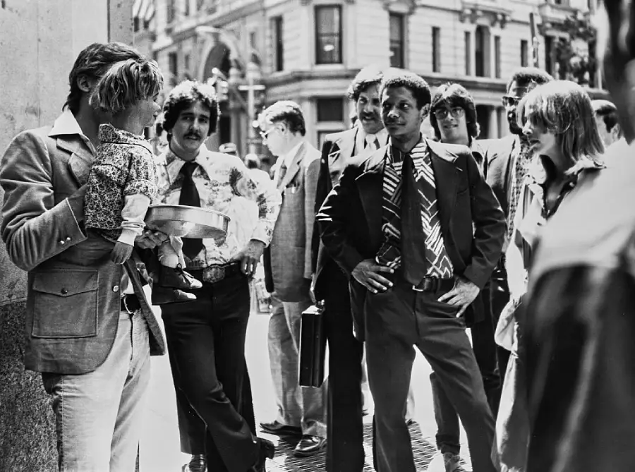 A group of people, some in suits, gather on a city street. One person holds a guitar and a doll. The mood appears lively as they interact and talk. Buildings and sunlight are visible in the background.