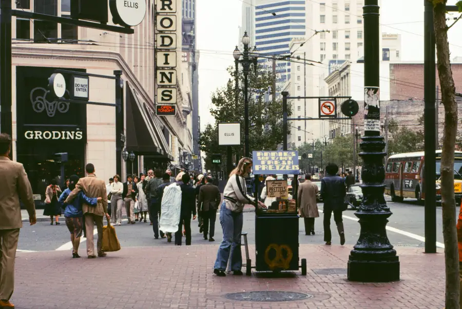 A city street scene with people walking, a pretzel stand in the foreground, tall buildings, a visible street sign for Ellis, and a Grodins store sign. A bus and streetlights are also visible in the background.
