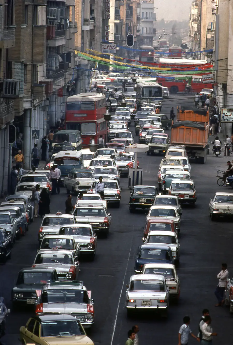 A busy city street packed with cars, trucks, and buses in heavy traffic. People walk along the sidewalks and some bicycles are present. Buildings line both sides of the street.
