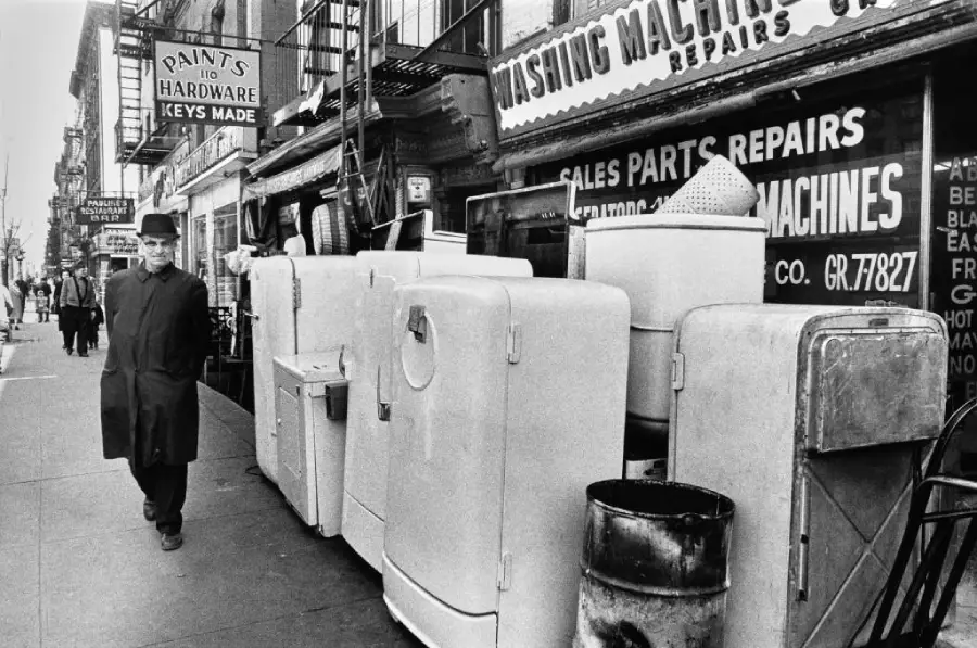Black and white photo of a man in a coat and hat walking past old refrigerators and appliances lined up on a city sidewalk in front of hardware and repair shops.