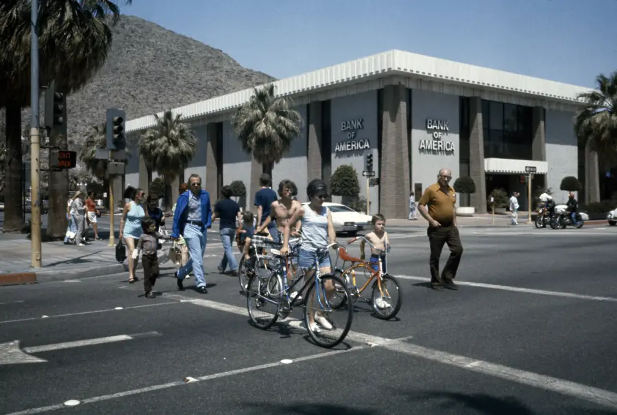 People, including children with bicycles, cross a sunny intersection near a Bank of America building with palm trees and a mountain in the background.