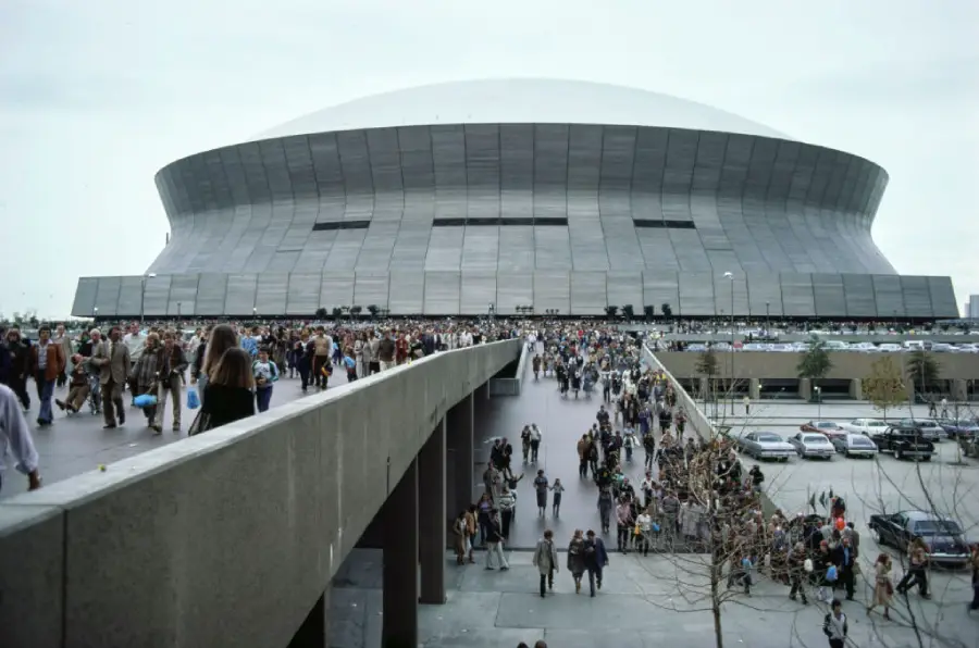 Crowds of people walk on a elevated ramp and gather outside a large, round stadium with a gray, domed roof on a cloudy day. Cars are parked in lots nearby.