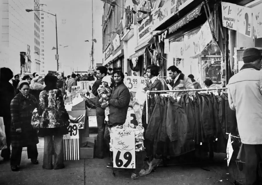 Black and white photo of a busy city street market with people shopping for clothes. Winter coats hang on racks and sale signs are visible. Shoppers and vendors interact on the crowded sidewalk.