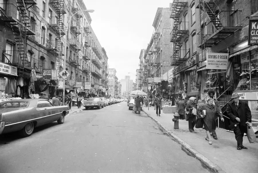 Black and white photo of a busy city street lined with parked cars, pedestrians, street vendors, and old buildings with fire escapes. Signs and storefronts are visible along both sides of the street.