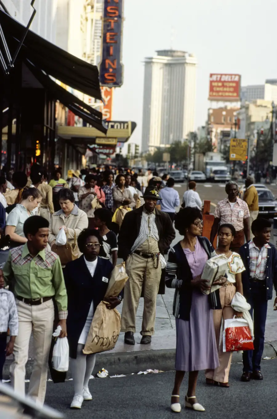 Crowd of people walking and shopping on a busy city street, carrying bags. Some look at storefronts or signs. Tall buildings, cars, and signs are visible in the background.