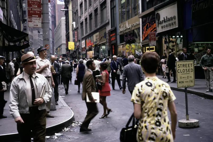 Busy city street in the 1960s or 70s with people walking, storefronts, a “Street Closed” sign, and various businesses visible in the background.
