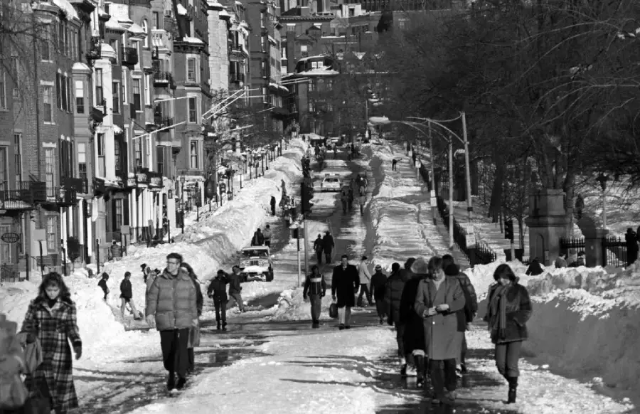 People walk along a snowy city street lined with buildings and parked cars. Snow is piled high on the sidewalks, and the scene appears cold and wintry.
