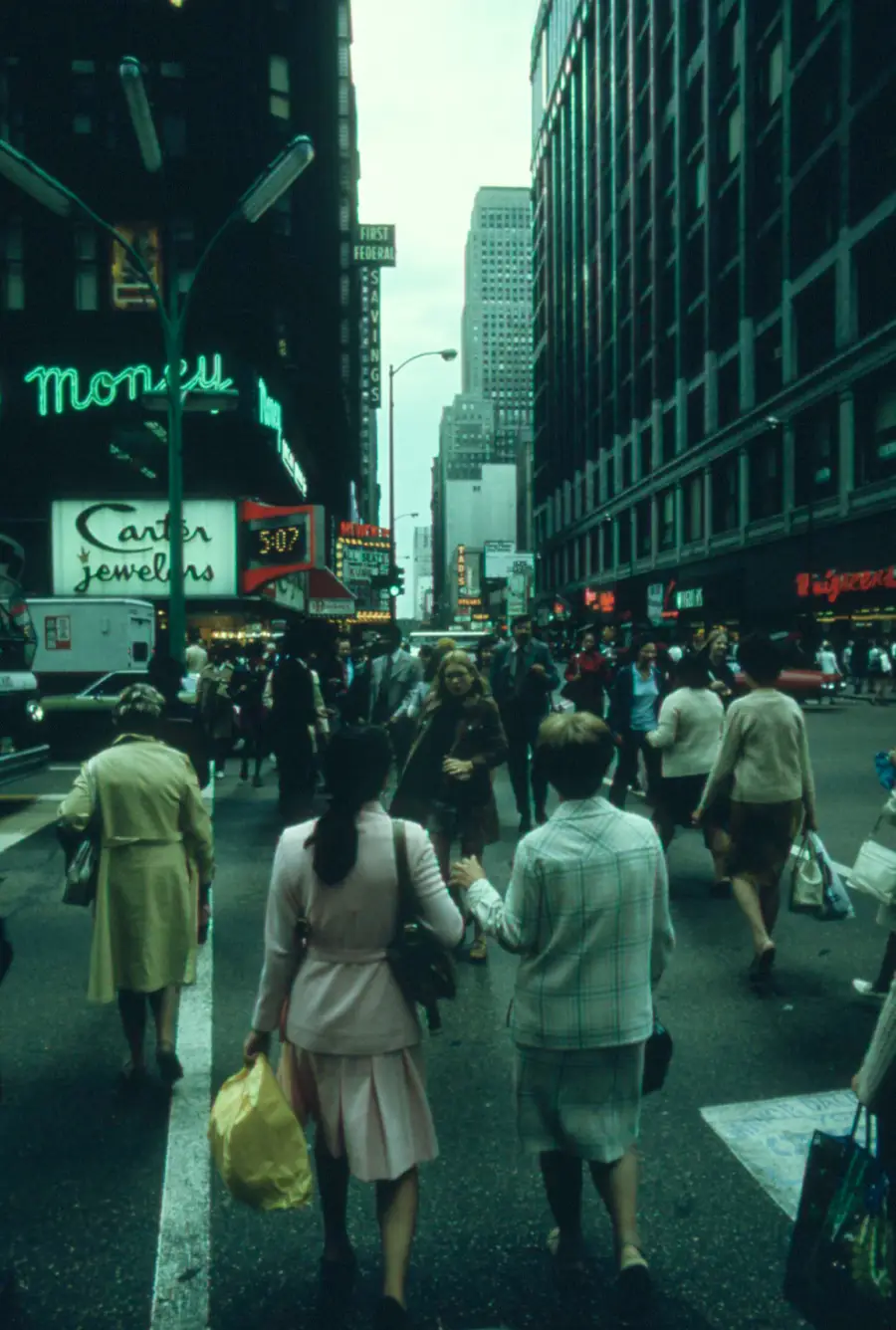People cross a busy city street surrounded by tall buildings, neon signs, and shops. Some carry shopping bags. The atmosphere suggests a bustling urban scene, likely from the past.