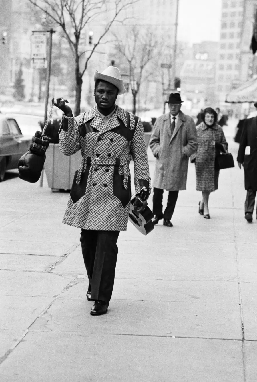 A man in a patterned coat carries boxing gloves and a bag while walking on a city sidewalk. Two people in winter coats walk behind him. Trees and buildings line the street.