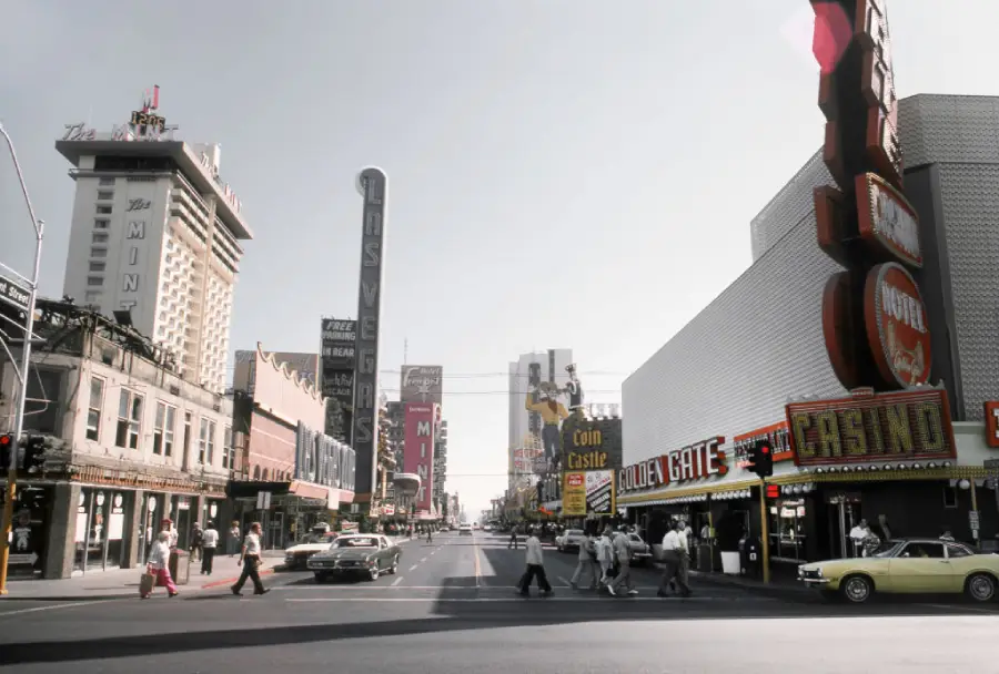 A vintage street scene of downtown Las Vegas featuring classic cars, people walking, and old casino signs like Golden Gate and Coin Castle under a clear sky.