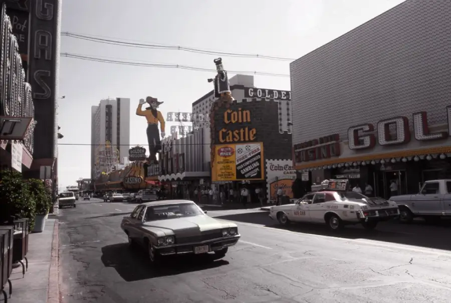 A vintage street scene shows casinos, neon signs, and classic cars in downtown Las Vegas. A cowboy sign stands above “Coin Castle” and “Golden Nugget” signs are visible, with people walking along the sidewalks.