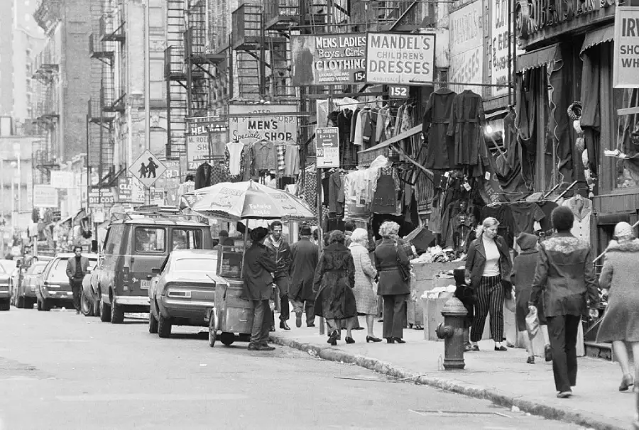Black-and-white photo of a busy city street with parked cars, people walking, street vendors, and buildings with clothing shops and fire escapes. Signs for dresses and clothing are visible.