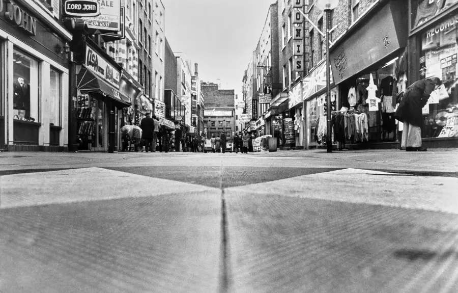 Black and white photo of a city street with shops on both sides, people walking, and the camera positioned low to the ground, capturing the scene from street level.