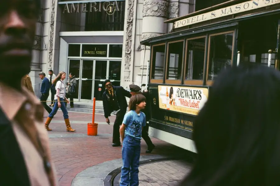 A boy stands near a cable car in a busy city street, with people walking by and ornate building details in the background.