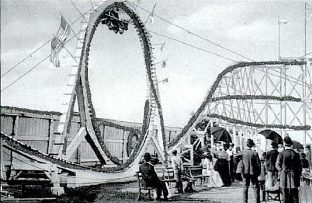 Black and white photo of an old-fashioned roller coaster with a large vertical loop, people standing and sitting nearby, and a group of riders upside down at the top of the loop.