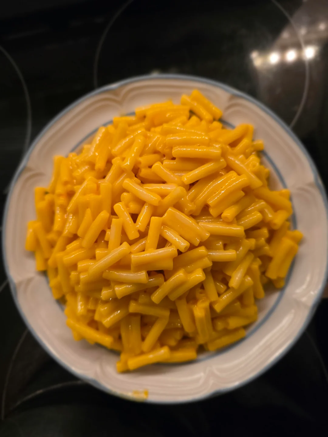 A plate piled high with cooked macaroni and cheese, featuring short, yellow pasta coated in creamy cheese sauce, sitting on a white plate with a blue rim.