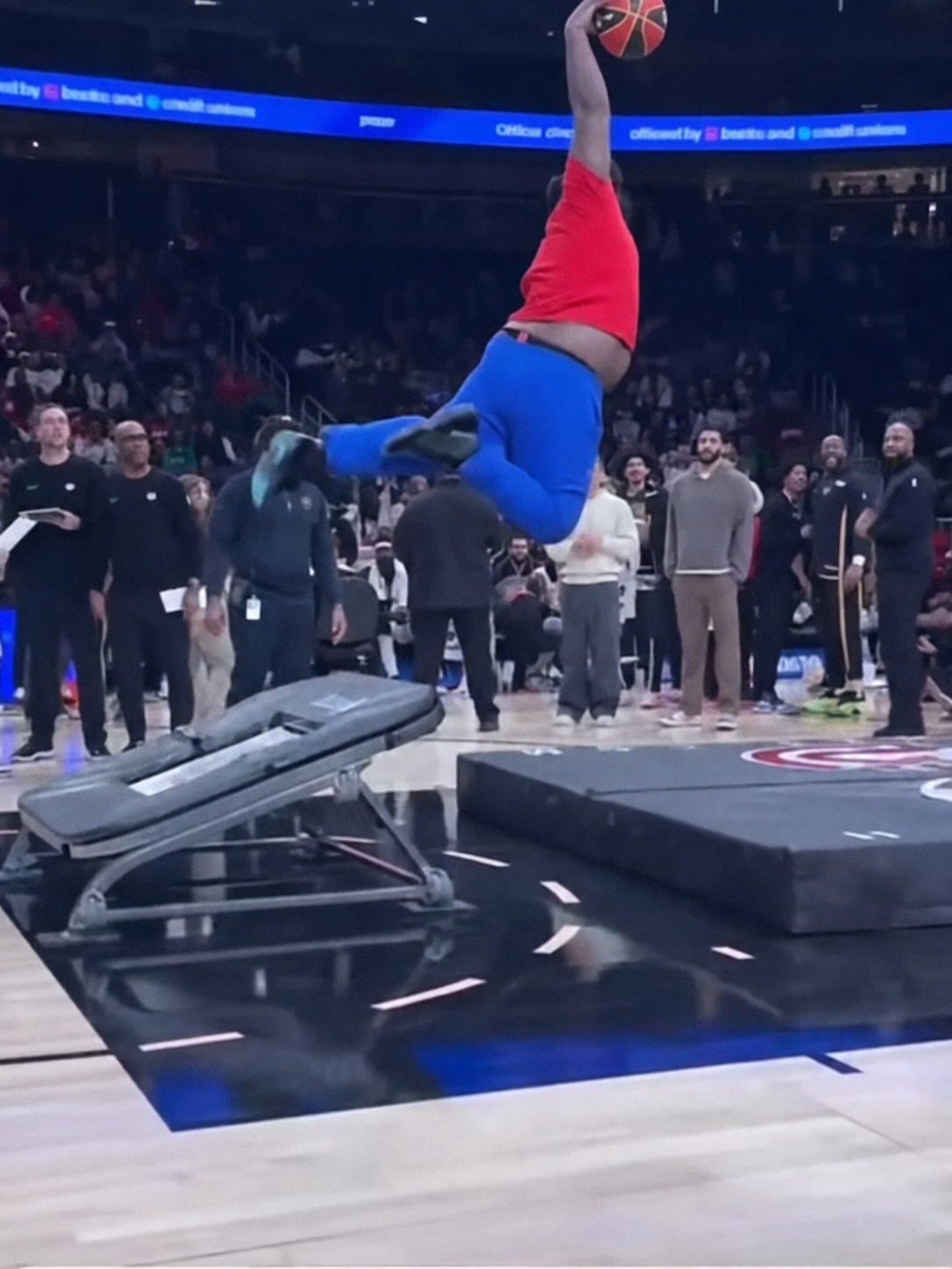 A performer in blue pants and a red shirt dunks a basketball after jumping off a trampoline, while a crowd watches in an indoor arena.