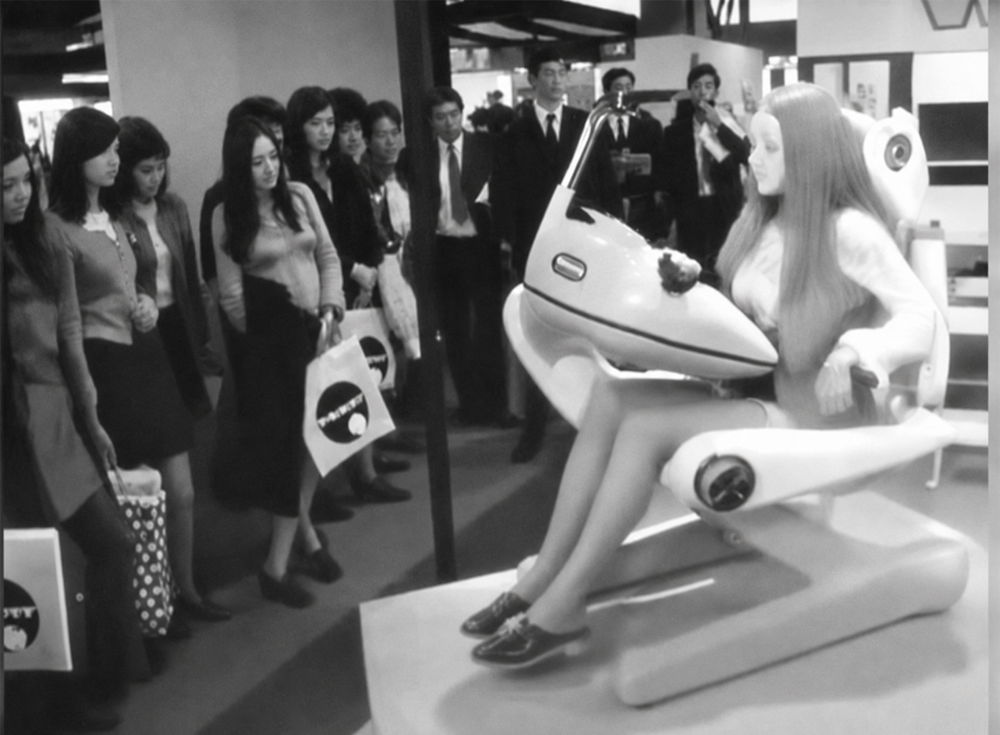 A group of people watches a seated mannequin with long hair using a futuristic machine at an indoor exhibition. Some people hold bags and appear intrigued by the display.