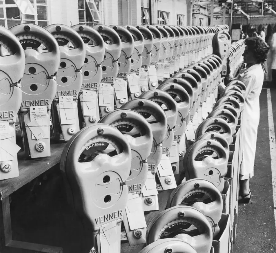 Rows of parking meters labeled “Venner” line a factory floor, with two women in white coats inspecting them. The image is black and white, and the meters are arranged in neat, orderly lines on shelves.