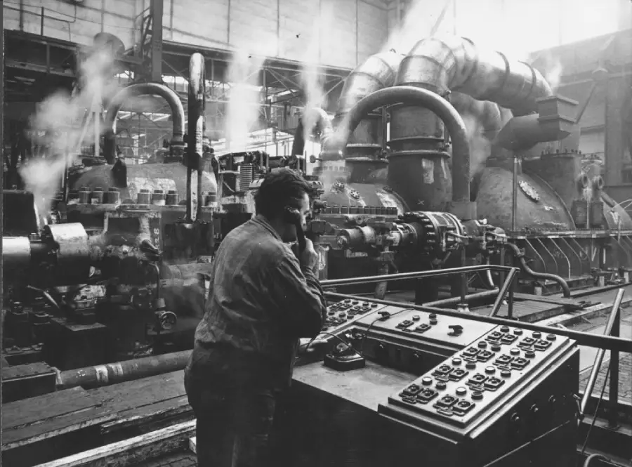 A worker operates a large control panel in an industrial factory with pipes, machinery, and steam in the background.