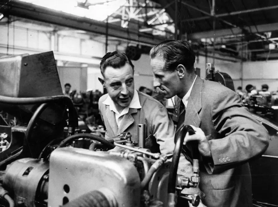 Two men in work attire examine machinery in a busy factory setting, engaged in conversation under industrial lighting, surrounded by metal equipment and tools.