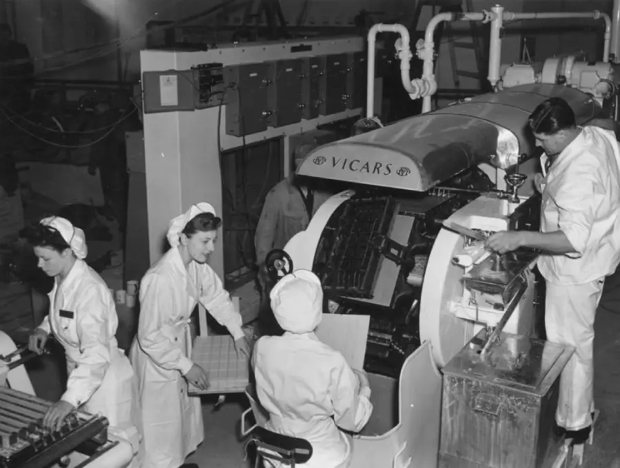 Four workers in uniforms and hair coverings operate a large Vicars machine in a factory, processing trays of items. Pipes, control panels, and tools are visible in the industrial setting. Black and white photo.