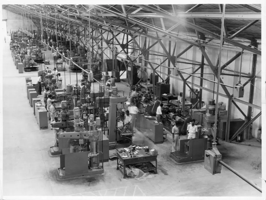 Black-and-white photo of a large industrial factory with rows of machinery and workers operating equipment along both sides of the building under a high, metal-framed roof.