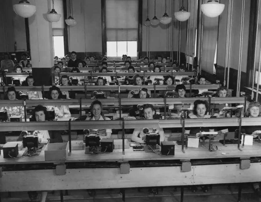 Black-and-white photo of women sitting in rows at desks in a large open room, working with telephones and switchboards, likely in a mid-20th-century telephone exchange.