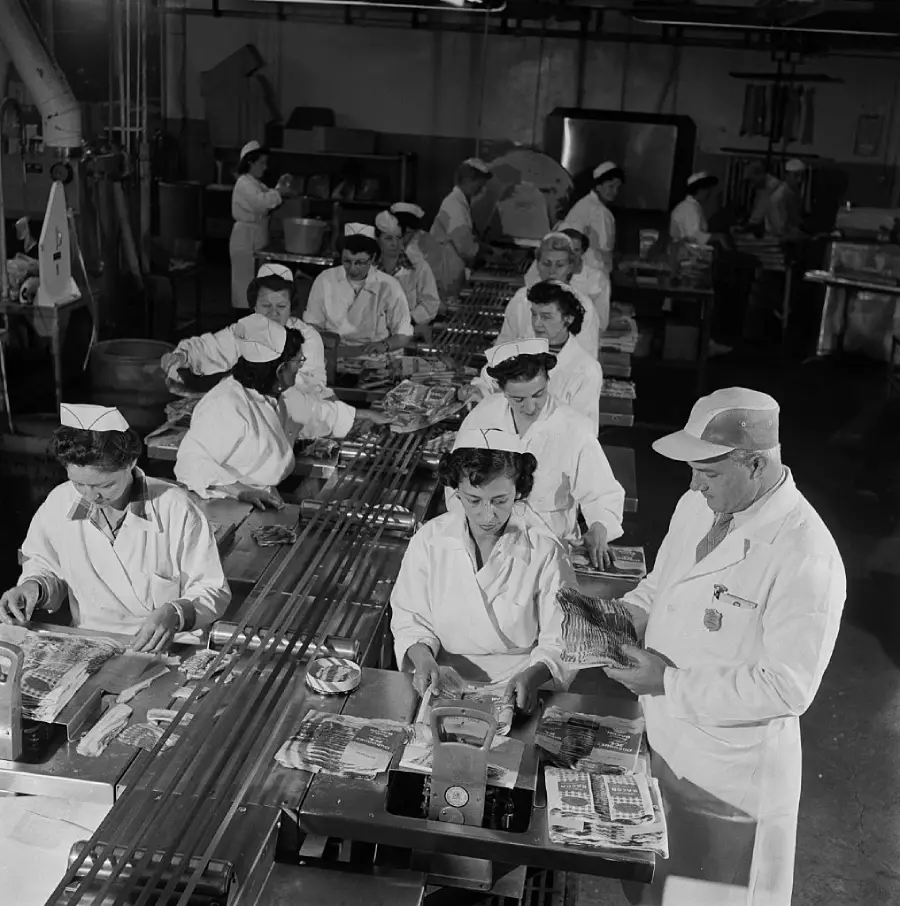 Workers in white uniforms and hats stand at a conveyor belt in a factory, packaging food products. The black-and-white image shows an assembly line process with several people focused on their tasks.