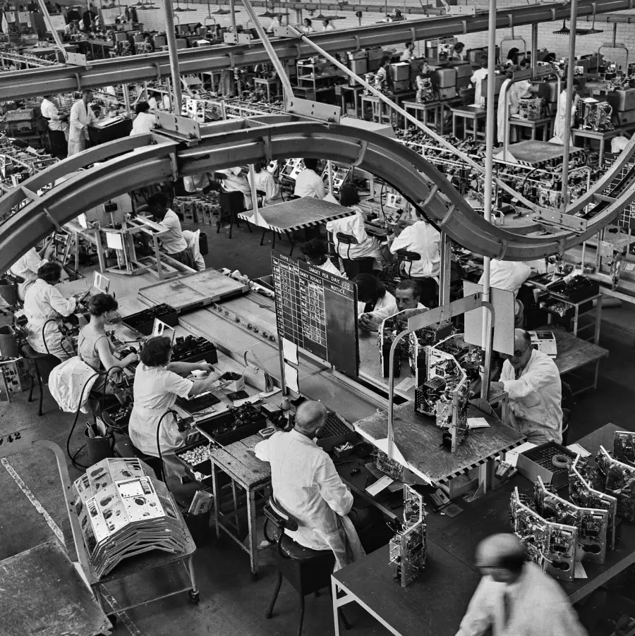Black and white photo of a busy electronics factory with workers in lab coats assembling devices at organized workstations, conveyor belts visible overhead.